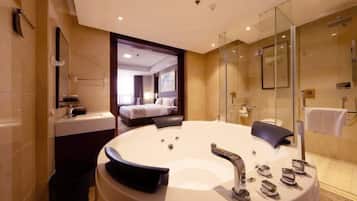 A modern bathroom with a large bathtub, a glass shower enclosure, and a visible bedroom with a bed and a window.