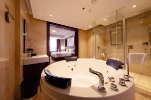Une salle de bain moderne dotée d’une grande baignoire, d’une cabine de douche en verre et d’une chambre visible avec un lit et une fenêtre.