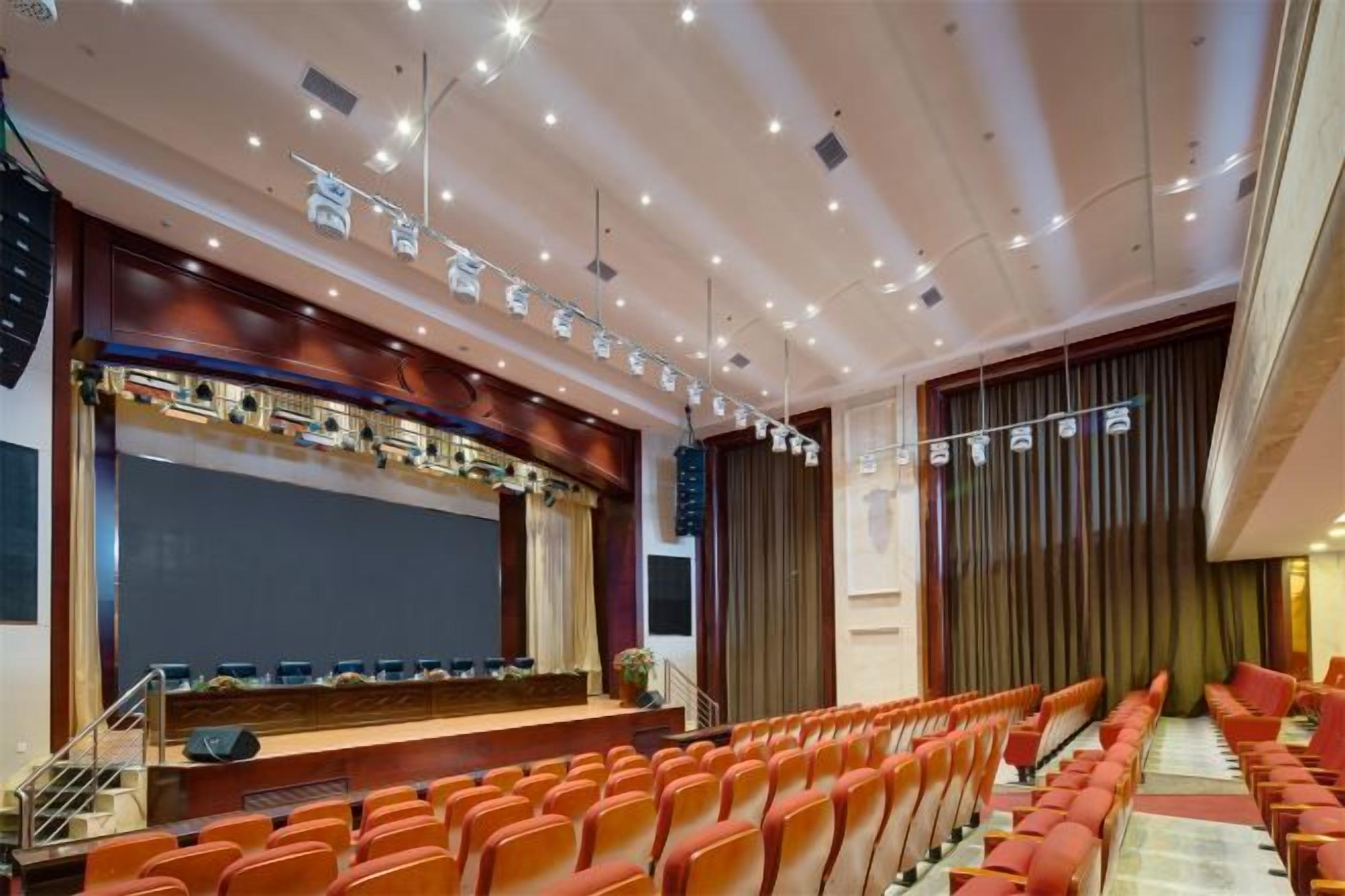 Uma sala de conferências com palco, fileiras de cadeiras e uma tela grande.