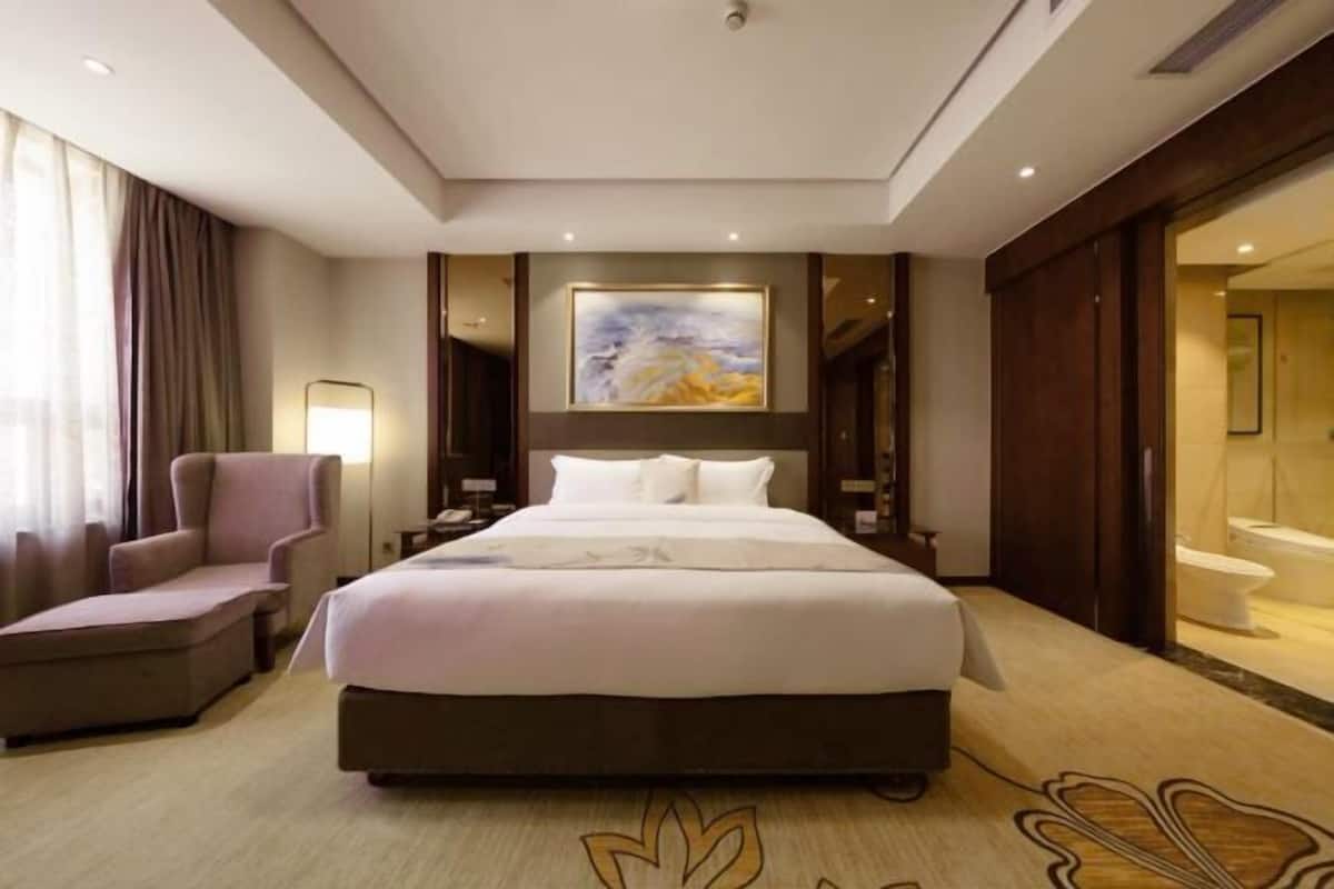 Une chambre d’hôtel avec un grand lit, une chaise, un bureau, un tableau accroché au mur et une salle de bain visible à travers une porte ouverte.