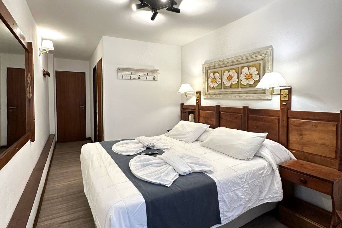 A hotel room with a large bed, wooden headboard, a nightstand, a bedside lamp, a wall-mounted artwork, and a hallway leading to another room.
