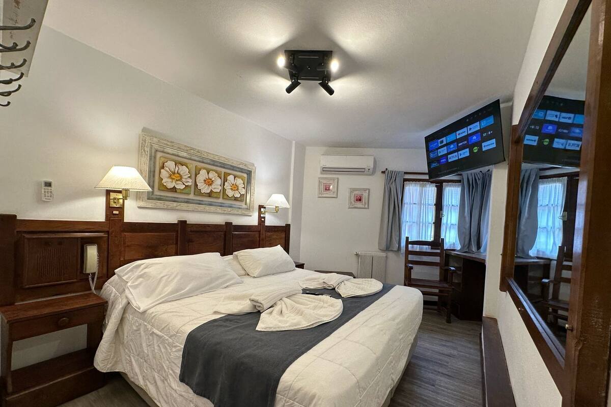 A hotel room with a bed, bedside lamps, a painting, an air conditioning unit, a television, and a desk with a chair.