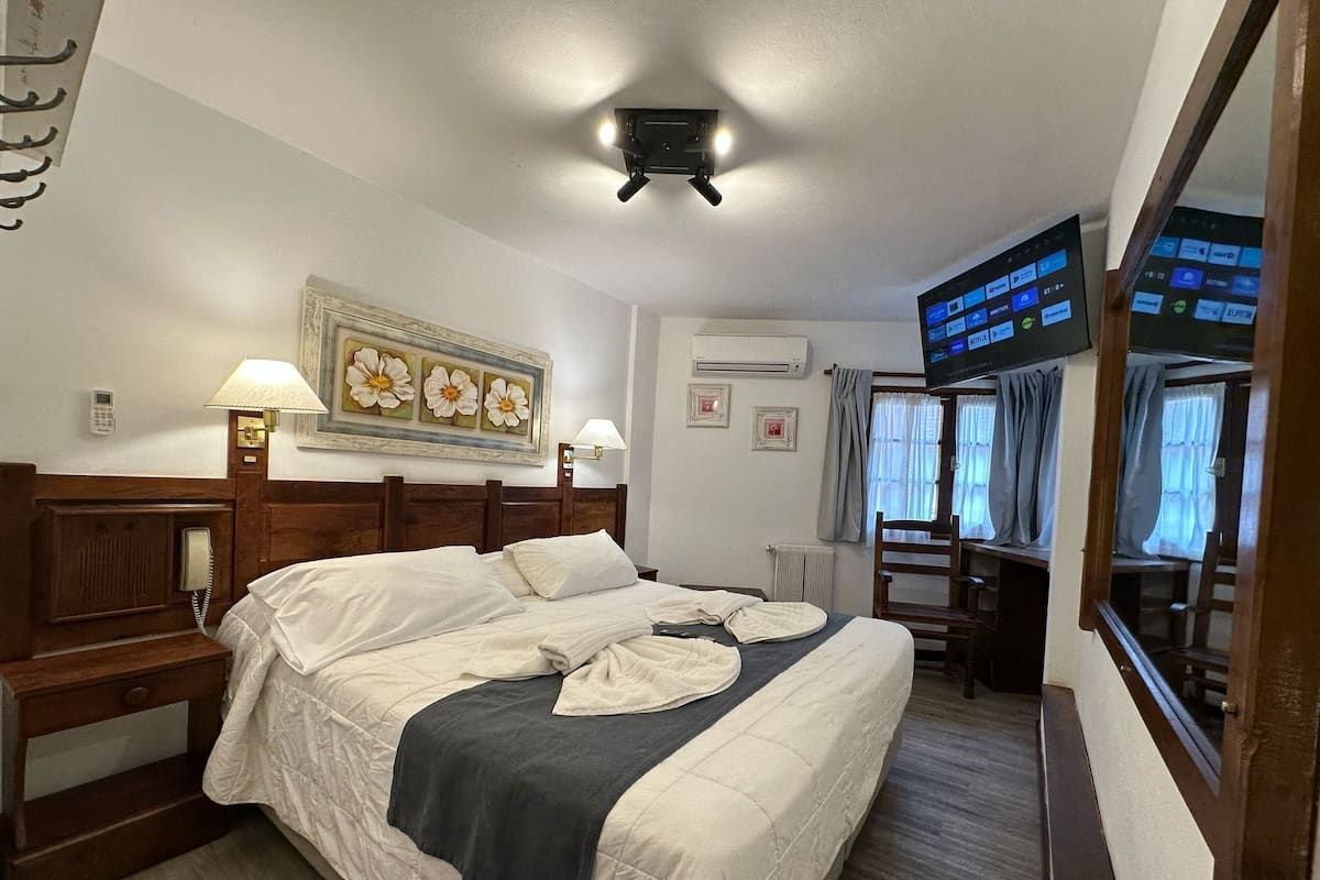 Habitación de hotel con cama, lámparas de noche, un cuadro, equipo de aire acondicionado, televisor y escritorio con silla.