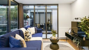 Un salon moderne avec un canapé bleu, un meuble TV noir et une grande fenêtre donnant sur l’extérieur.