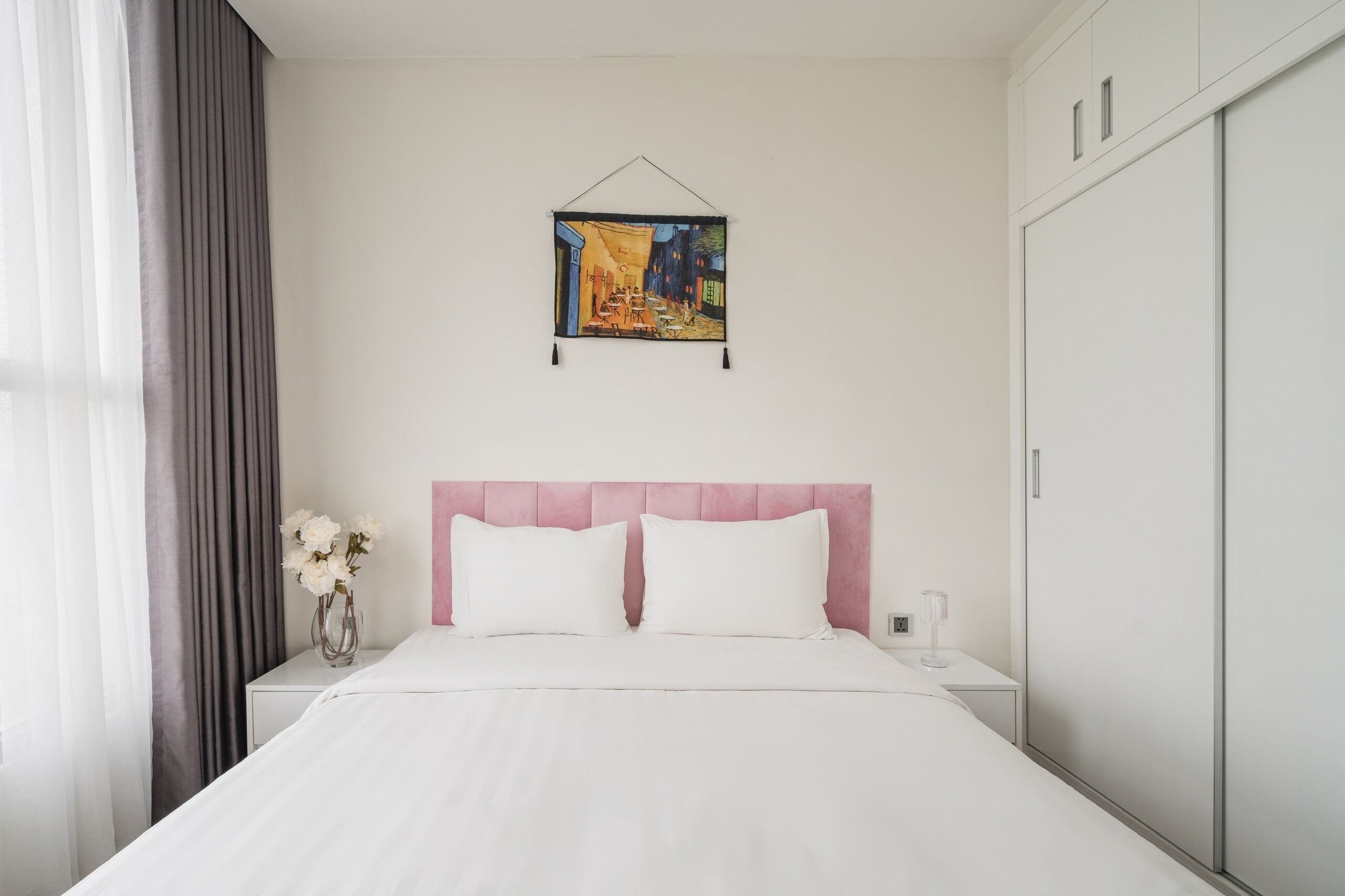 A neatly made bed with white linens, a pink headboard, and a wall-mounted painting.