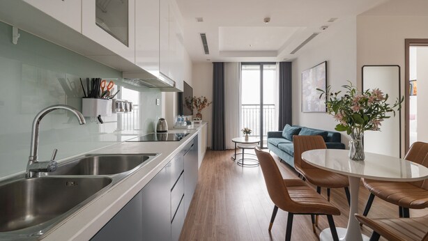 A modern kitchen with stainless steel appliances, a dining area with a round table and leather chairs, and a living room with a sofa and a large window.