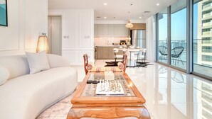 Uma sala de estar moderna com um sofá branco, uma mesa de centro de madeira com peças de xadrez, e vista para a cozinha e varanda.