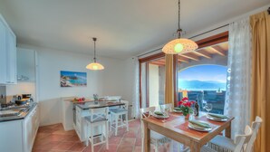 A dining area with a wooden table set for four, white chairs, and a view of the mountains through a large window. - Borgo Belvedere Apartment With Lake View (Toscolano Maderno)