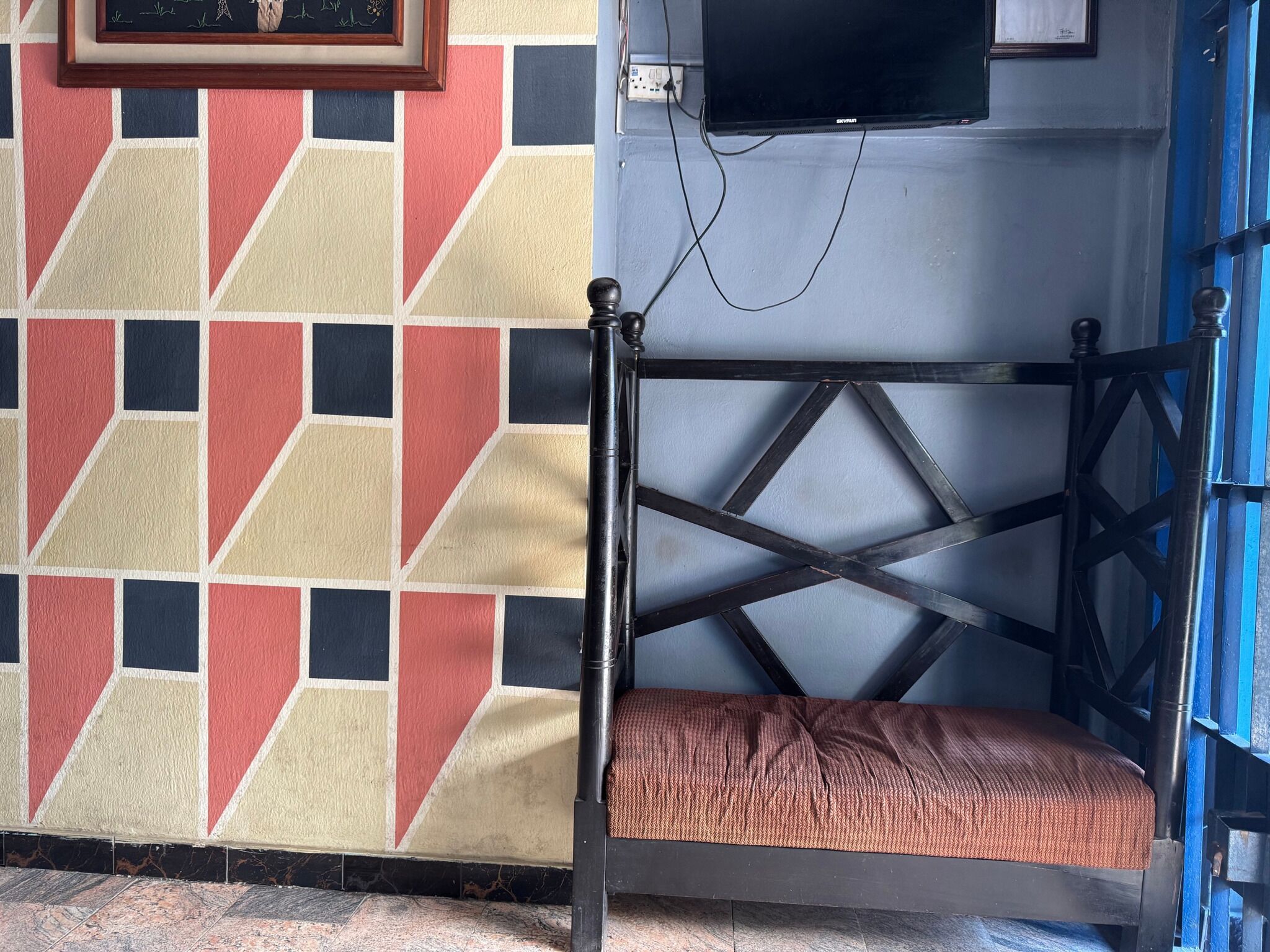 Un coin de pièce avec un mur orné d’un motif géométrique, un banc à ossature métallique et une porte bleue.