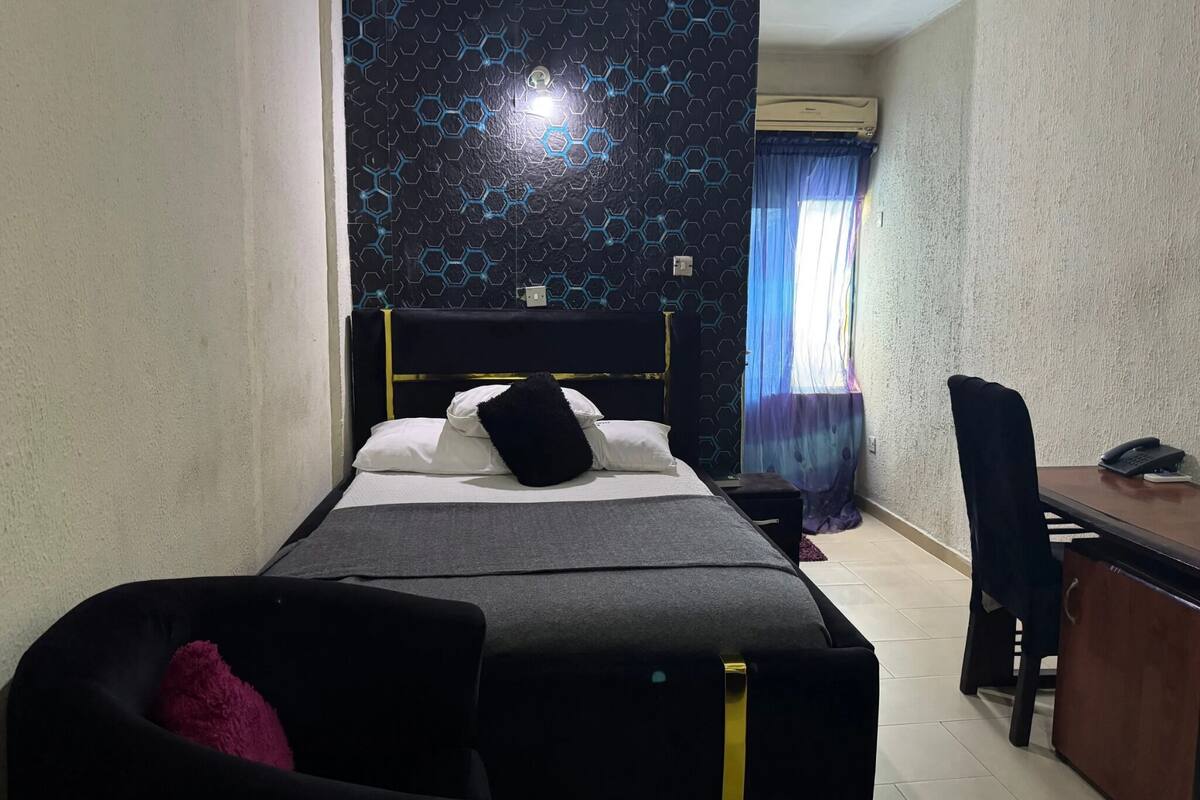 A hotel room with a bed, a desk, a chair, and an air conditioning unit.