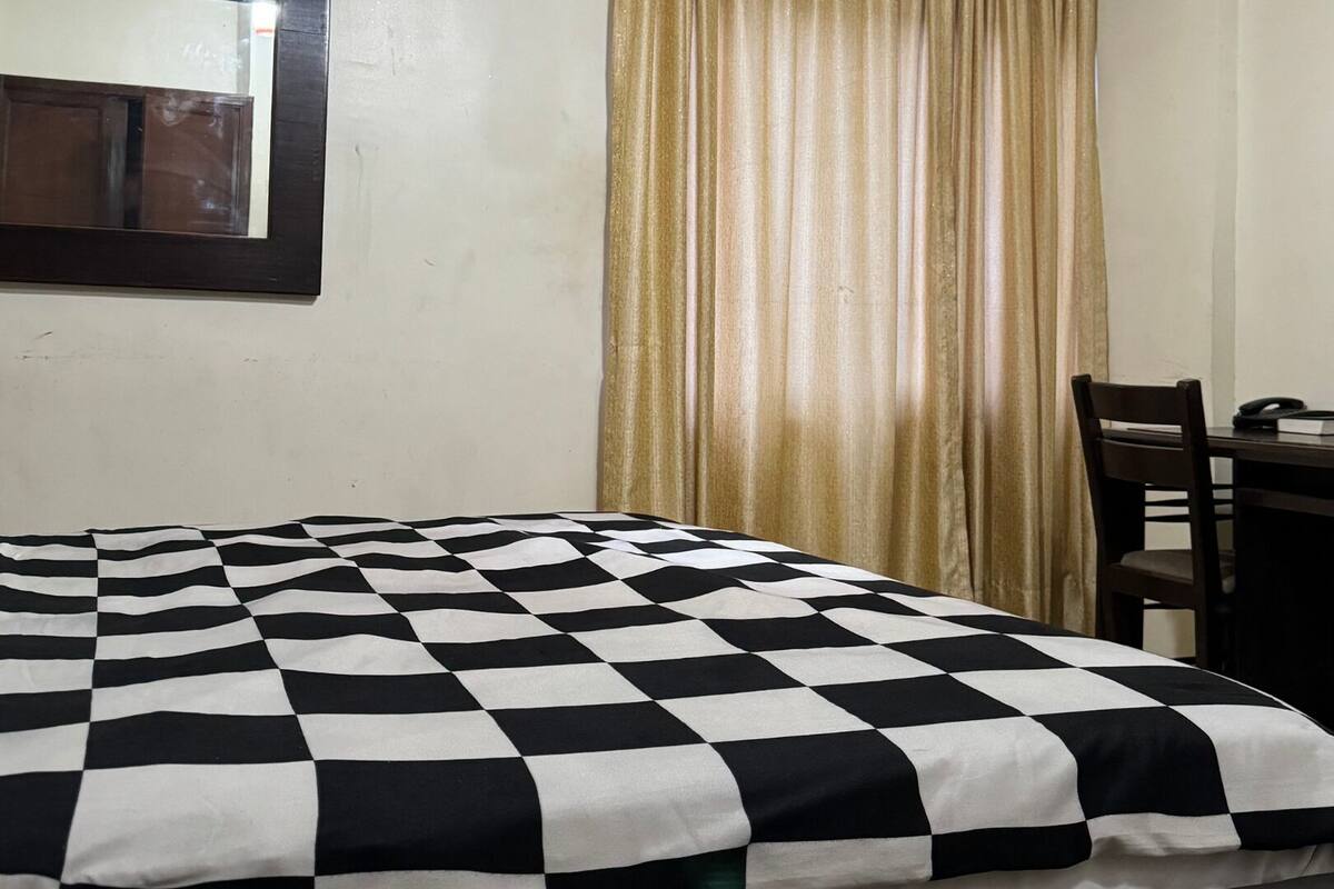 A room with a bed covered in a checkered blanket, a desk with a chair, a television on the wall, and a window with curtains.