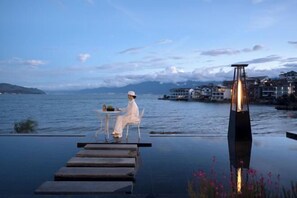 Une personne est assise à une table au bord d’une piscine à débordement, avec une vue sur une étendue d’eau et des bâtiments en arrière-plan.