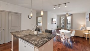 A modern kitchen with a marble countertop, stainless steel sink, pendant lighting, and a dining area with white chairs and a table.