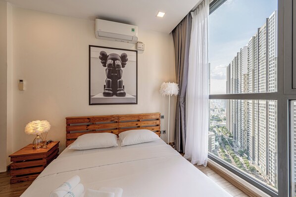 Habitación de hotel con una cama grande, cabecera de madera, un cuadro en la pared, aire acondicionado, mesita de noche con lámpara y vista a edificios altos a través de una ventana.