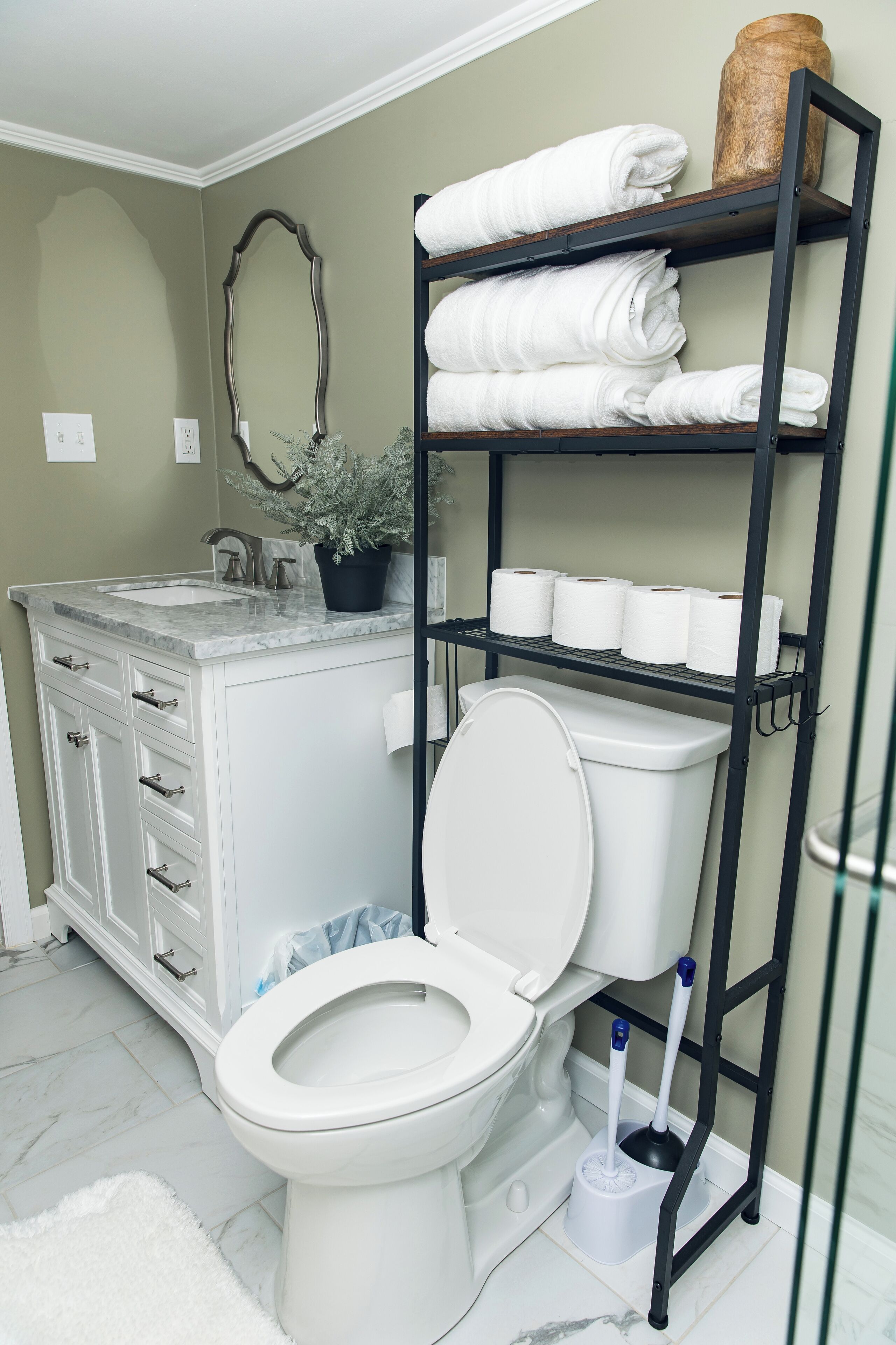A bathroom with a toilet, a sink, a marble countertop, a white cabinet with drawers, a shelf with towels, and a toilet brush.