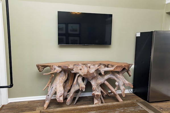 Una habitación con una mesa de madera, un televisor de pantalla plana en la pared y un refrigerador.