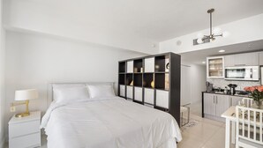 Et moderne soveværelse med en stor seng, et natbord med lampe, en indbygget reol og et tekøkken i baggrunden.