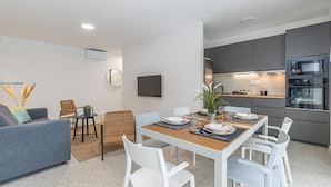 A modern living room with a dining table, white chairs, a grey sofa, and a kitchen area with grey cabinets and a built-in oven. - Pool Apartment Sapore Di Mare 3 (Baska)