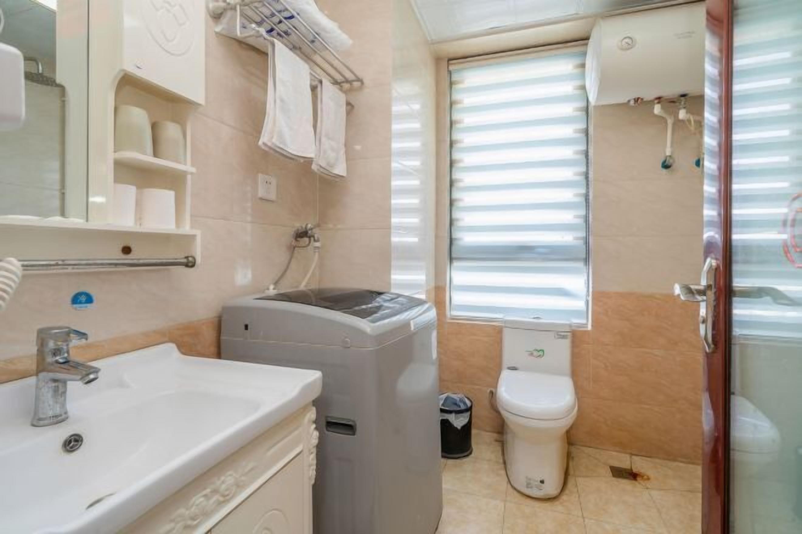 A bathroom with a sink, a washing machine, a toilet, and a window with blinds.