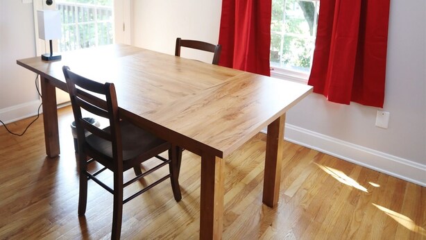 Une salle à manger avec une table en bois, deux chaises, un lustre et une fenêtre ornée de rideaux rouges.