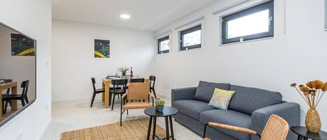 A modern living room with a grey sofa, wicker chairs, a small round table, and a dining area with a wooden table and chairs.