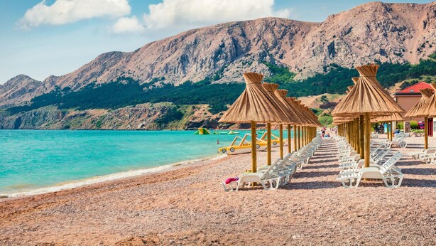 A beach with sun loungers and thatched umbrellas, a rocky mountain backdrop, and clear turquoise water. - Pool Apartment Sapore Di Mare 4 (Baska)