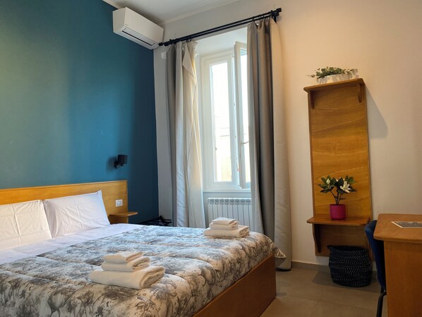 Un dormitorio con cama, mesita de noche, aire acondicionado montado en la pared, ventana con cortinas, estante de madera con una planta y un cubo de basura.