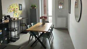 Una cucina moderna con un tavolo di legno, sedie e un frigorifero. Si vede una finestra con persiane, una pianta in vaso e uno specchio a parete.