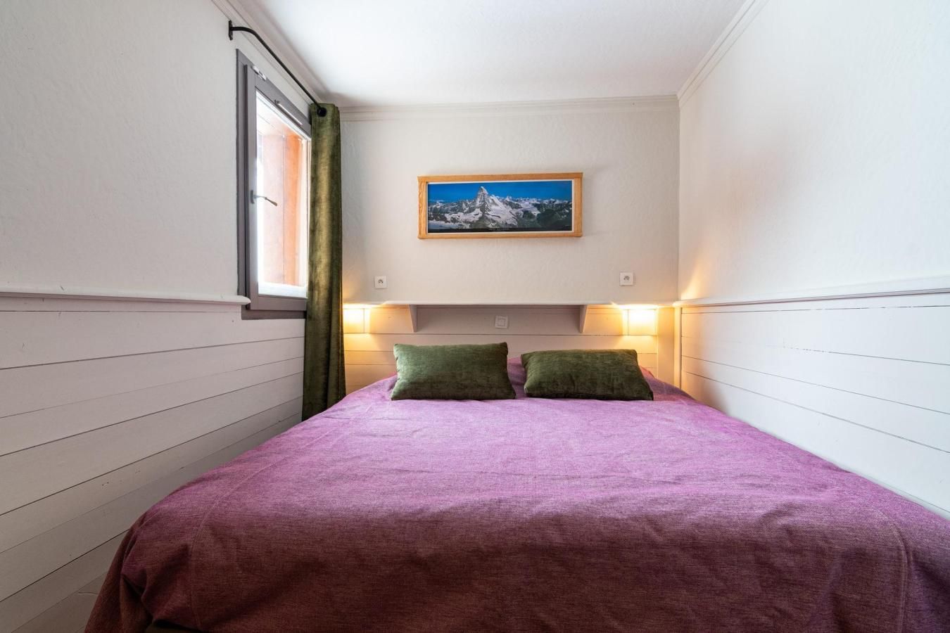 Een kleine kamer met een bed, een raam met gordijnen en een ingelijste foto aan de muur.