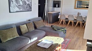 A modern living room with a grey sofa, a coffee table, a patterned rug, a dining area with chairs, a kitchen with a fridge, and a mountain landscape painting. - 100m from ski slopes, 3rd floor, swimming pool, sauna, hammam, balcony, fireplace or stove, tv (Val Thorens)