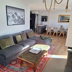 Una sala de estar moderna con un sofá gris, una mesa de centro, un tapete estampado, una zona de comedor con sillas, una cocina con refrigerador y una pintura de paisaje de montaña.