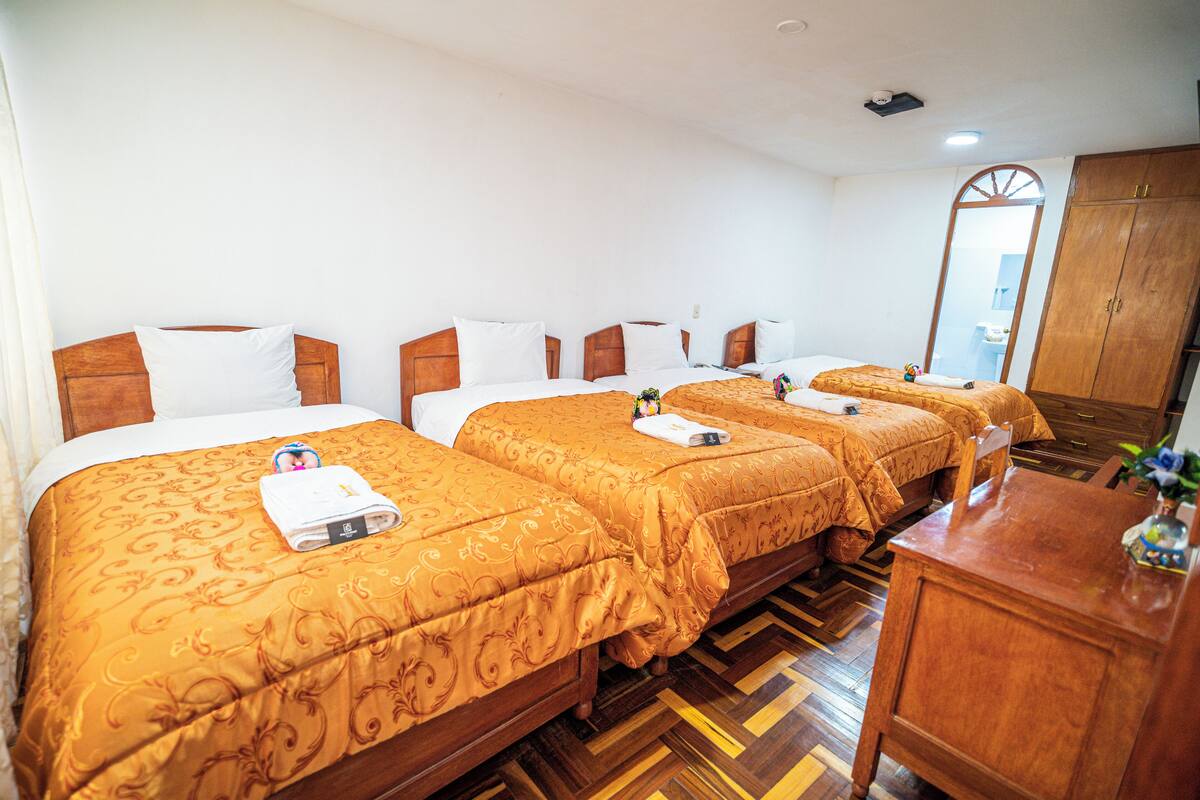 Une chambre d’hôtel avec quatre lits, chacun doté d’une tête de lit en bois et de draps blancs. On y trouve également une commode en bois et un miroir accroché au mur.
