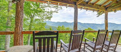 Una terraza de madera con mecedoras y una mesa, con vistas a un paisaje montañoso.