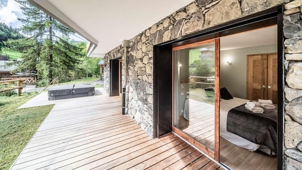 Une maison moderne avec une façade en pierre, une terrasse en bois et une grande porte-fenêtre coulissante donnant sur une chambre à coucher équipée d’un lit et d’une armoire en bois.