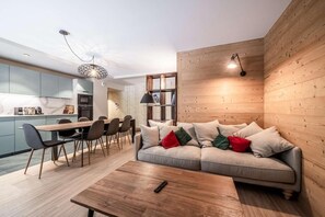 A modern living room with a wooden wall, a sofa with cushions, a dining area with chairs, and a kitchen in the background.