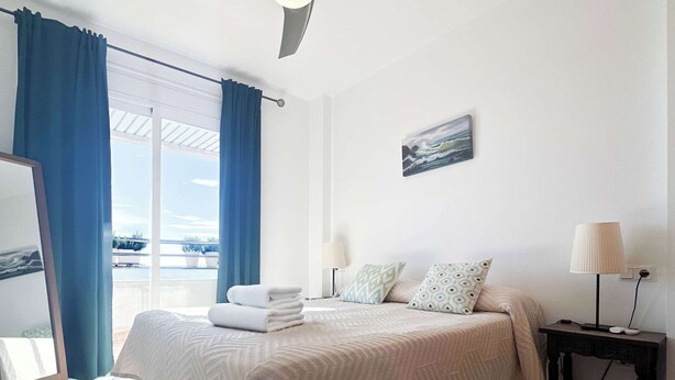 A hotel room with a bed, a ceiling fan, a nightstand, a lamp, a painting, and a view of the sea through a window. - Apartment Aguamarina 4B (Estepona)