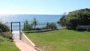 A grassy area with a metal gate leading to a body of water, surrounded by greenery and a bench. - Adosado junto al Mar (Estepona)