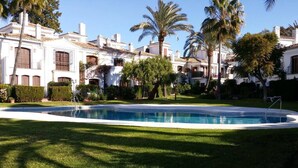 A swimming pool surrounded by well-maintained lawns and palm trees, with a building featuring traditional architecture in the background. - Adosado junto al Mar (Estepona)