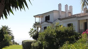 A white building with a balcony, surrounded by lush greenery and palm trees. - Adosado junto al Mar (Estepona)