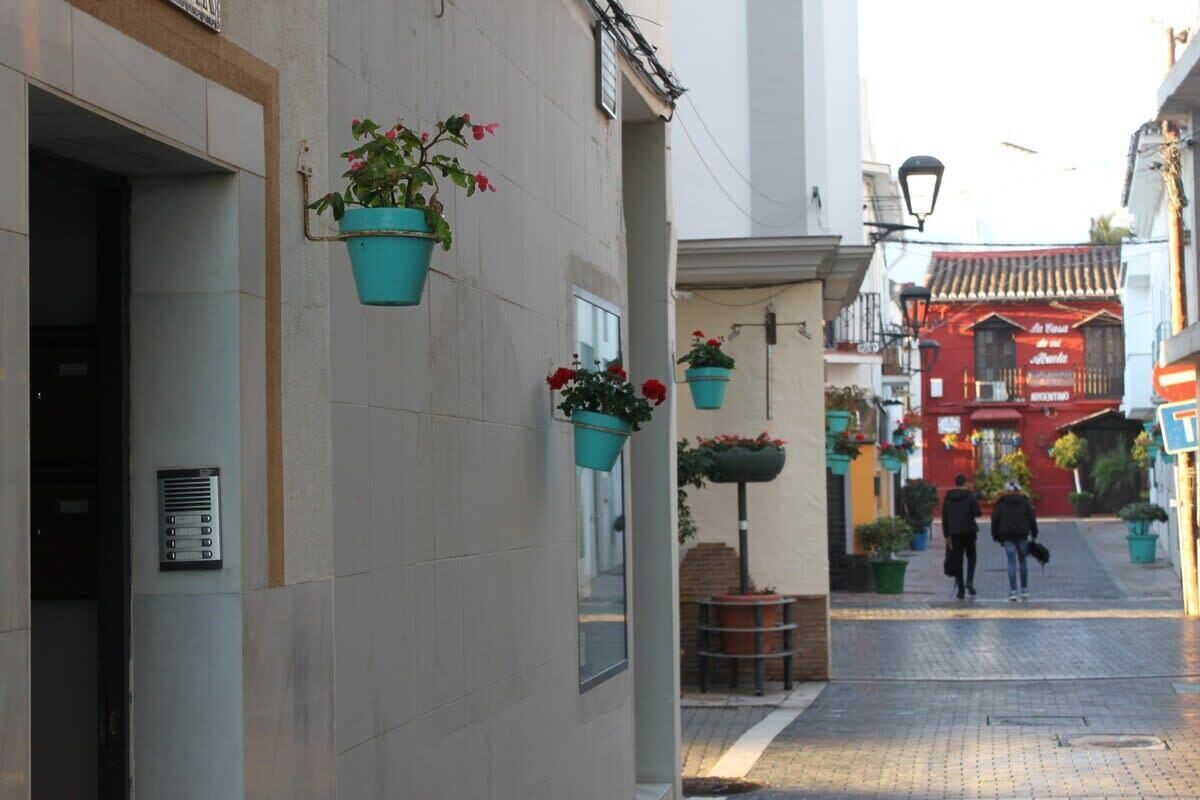Une rue étroite, ornée de pots de fleurs suspendus, avec un bâtiment rouge en arrière-plan.