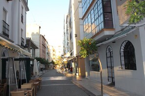 A narrow street lined with buildings, some with balconies and others with large windows. There are outdoor seating areas with tables and chairs.
