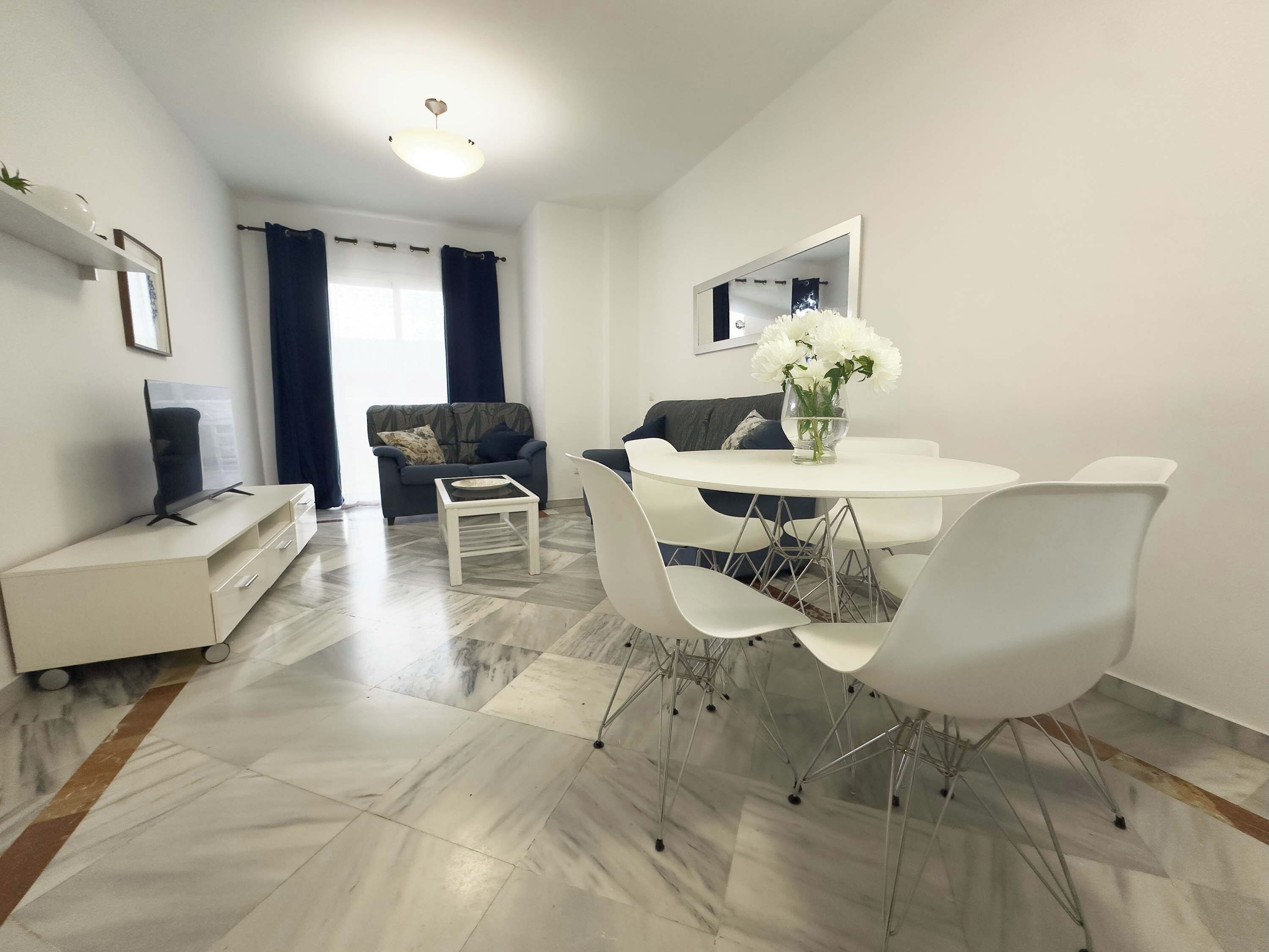 A modern living room with a sofa, a dining table with chairs, a TV, and a vase with flowers.
