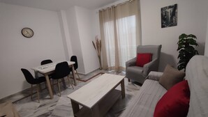 Uma sala de estar com sofá, mesa de jantar com cadeiras, mesa de centro, poltrona cinza com almofada vermelha, planta em vaso, relógio de parede e janela com cortinas.