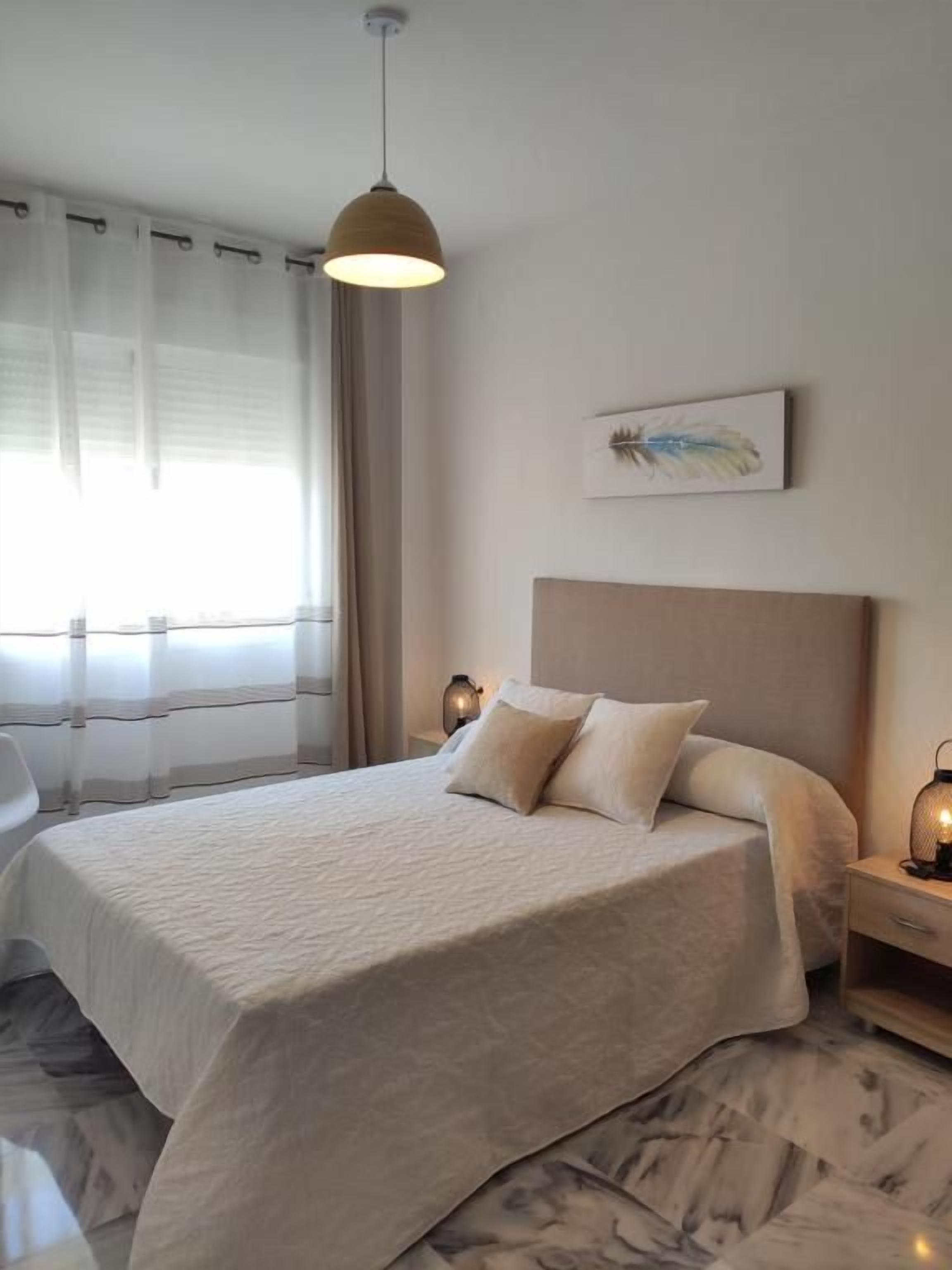 A modern bedroom with a large bed, a bedside table with a lamp, and a window with sheer curtains.