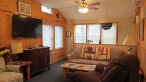 L’intérieur d’un chalet accueillant, doté d’un téléviseur, de murs en bois, d’un ventilateur de plafond et d’une fenêtre avec des stores.