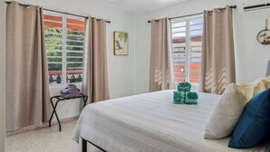 A bedroom with a bed, curtains, a window with a view, a small table with towels, and a wall-mounted air conditioner. - Tropical Oasis Retreat & Infiniti Pool (Luquillo)
