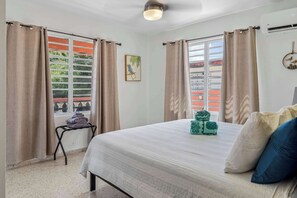 A bedroom with a bed, curtains, a window with a view, a small table with towels, and a wall-mounted air conditioner.