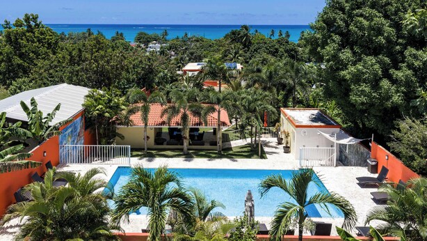 A pool area with a clear view of the ocean, surrounded by lush greenery and a building with a red roof. - Tropical Oasis Retreat & Infiniti Pool (Luquillo)