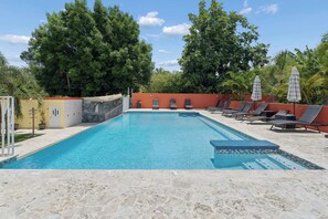 A swimming pool with lounge chairs and umbrellas, surrounded by trees and a stone wall.