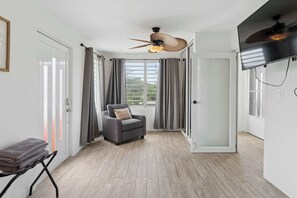 A room with a grey sofa, a standing fan, a TV, and a window with curtains.